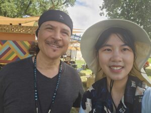 Menominee rock artist Wade Fernandez and WFU Journalism minor Hope Zhu stop for a picture during the Smithsonian Folklife Festival. They are both smiling in front of a backdrop of colorful stands, trees, and a partly cloudy sky. 