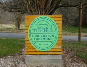 This photograph shows the sign, a bright green and blue logo reading "Our Boston Thurmond" on a wood paneled background, recognizing the historic community.